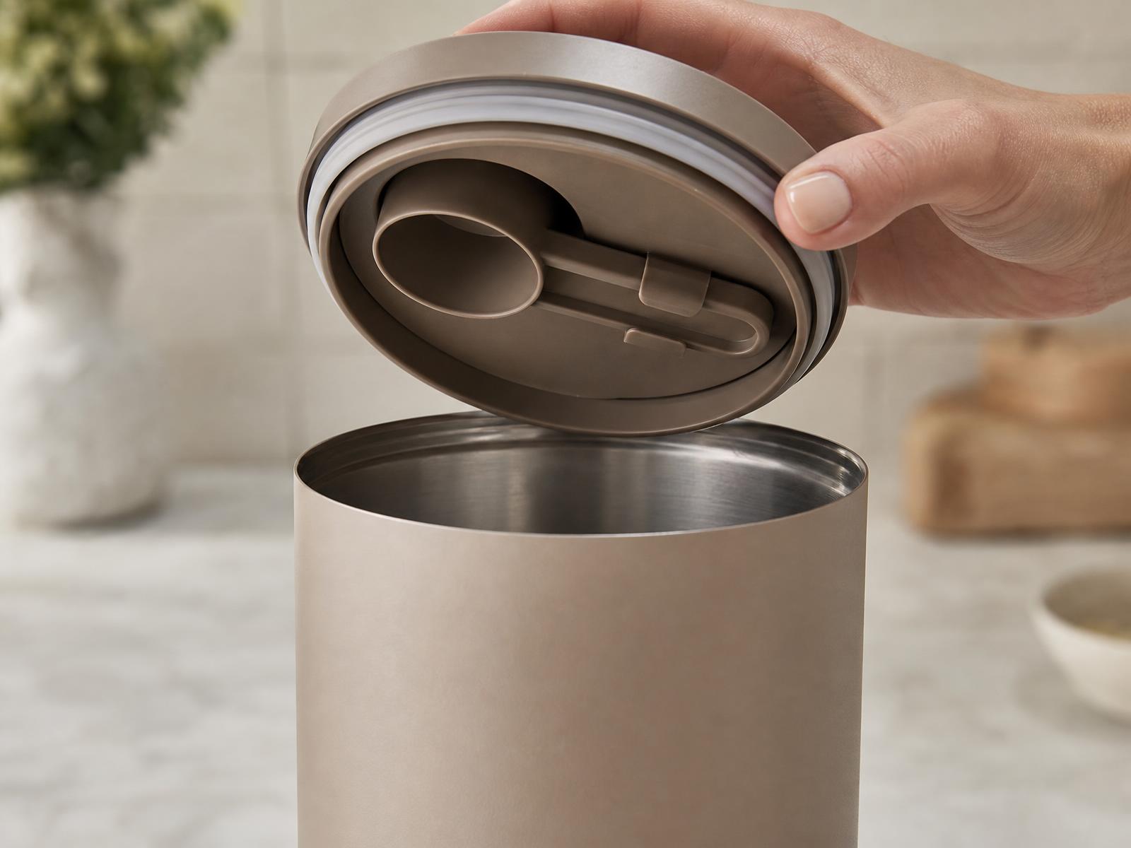Lid lifted off a stainless canister showing the measuring scoop clipped into the underside of the lid